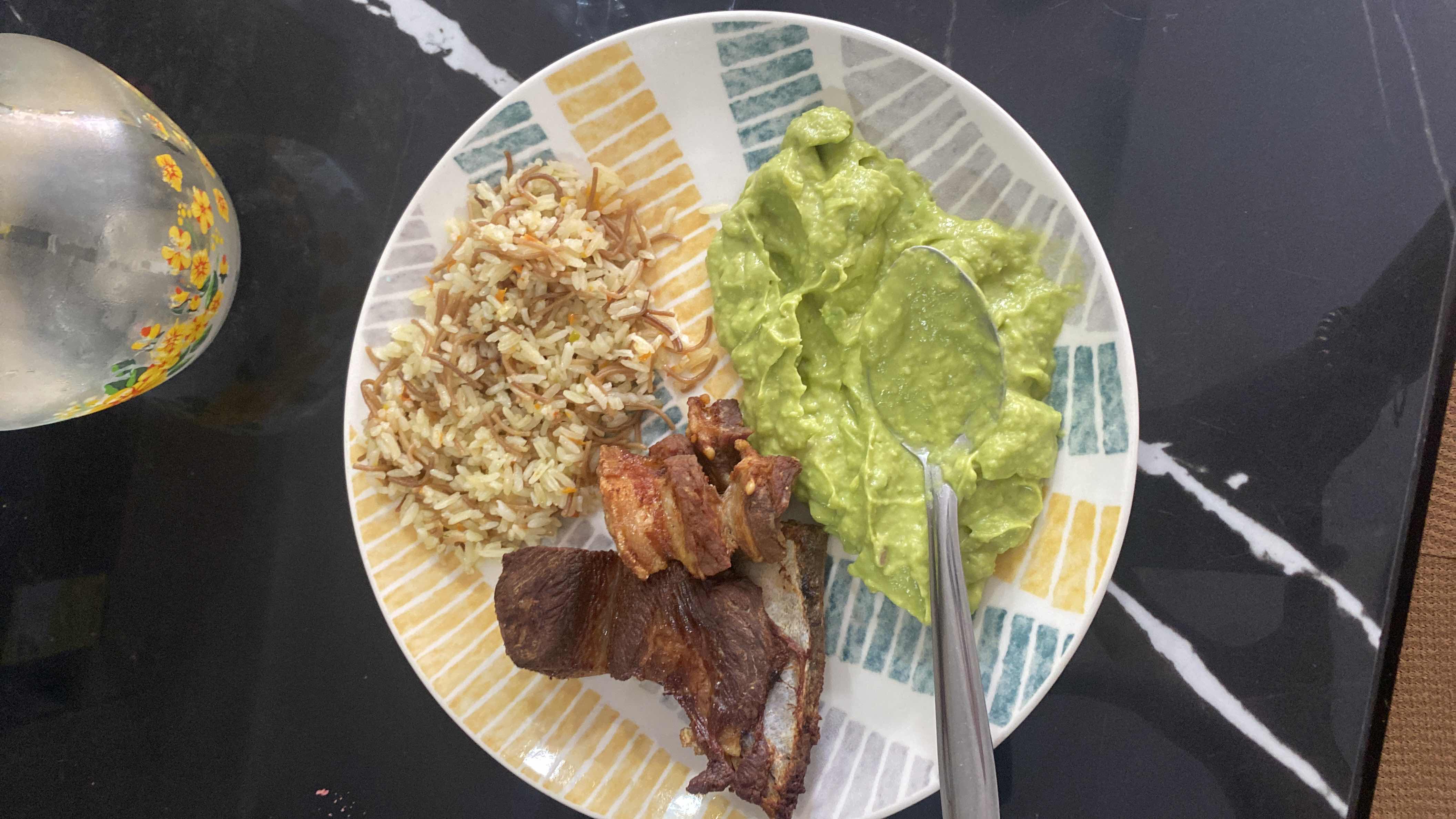A plate containing rice with vermicelli, grilled meat, and mashed avocado, providing a balanced mix of protein, carbs, and fats.