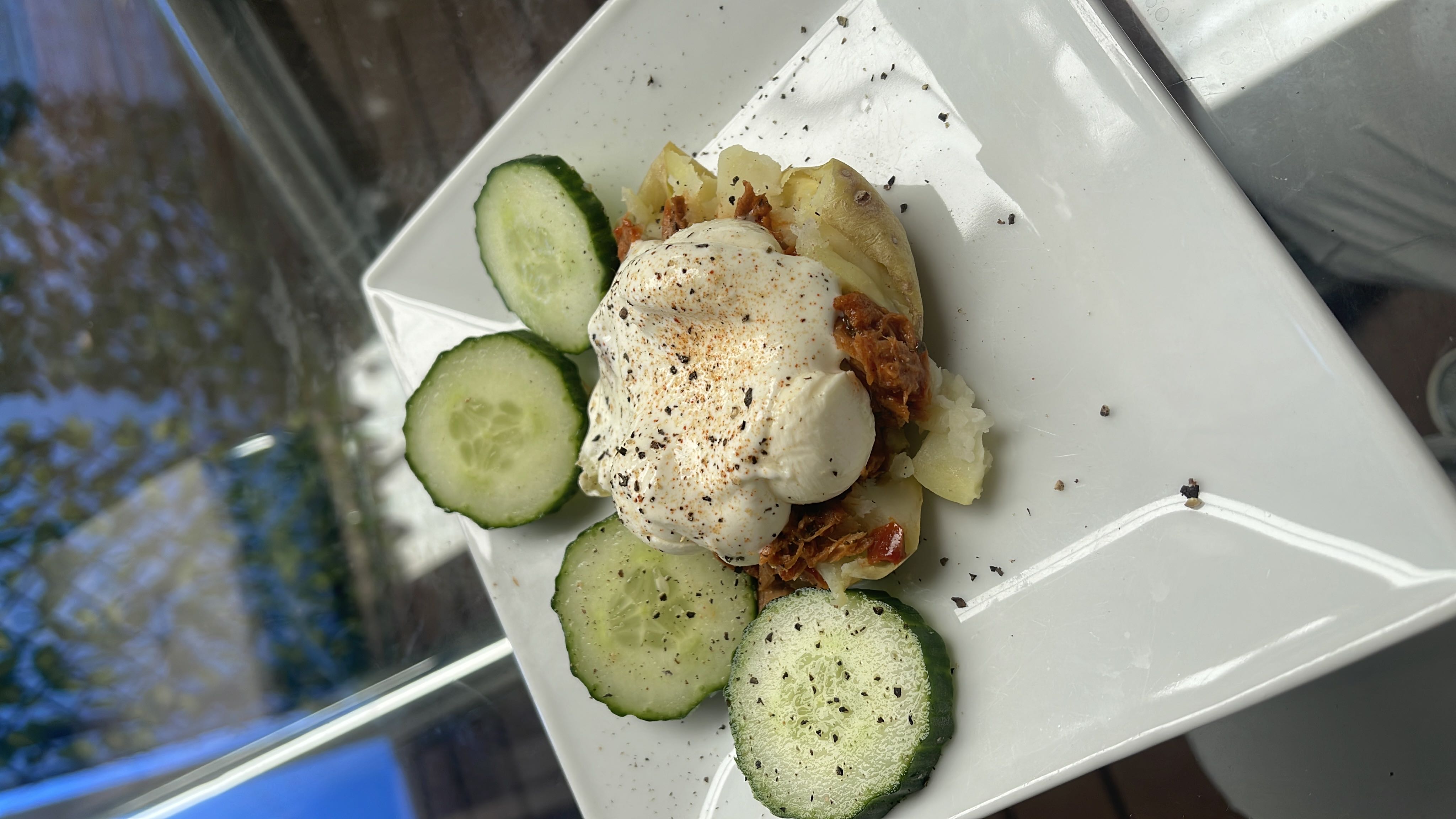 A dish featuring a baked potato topped with pulled meat and sour cream, accompanied by cucumber slices, seasoned with black pepper.