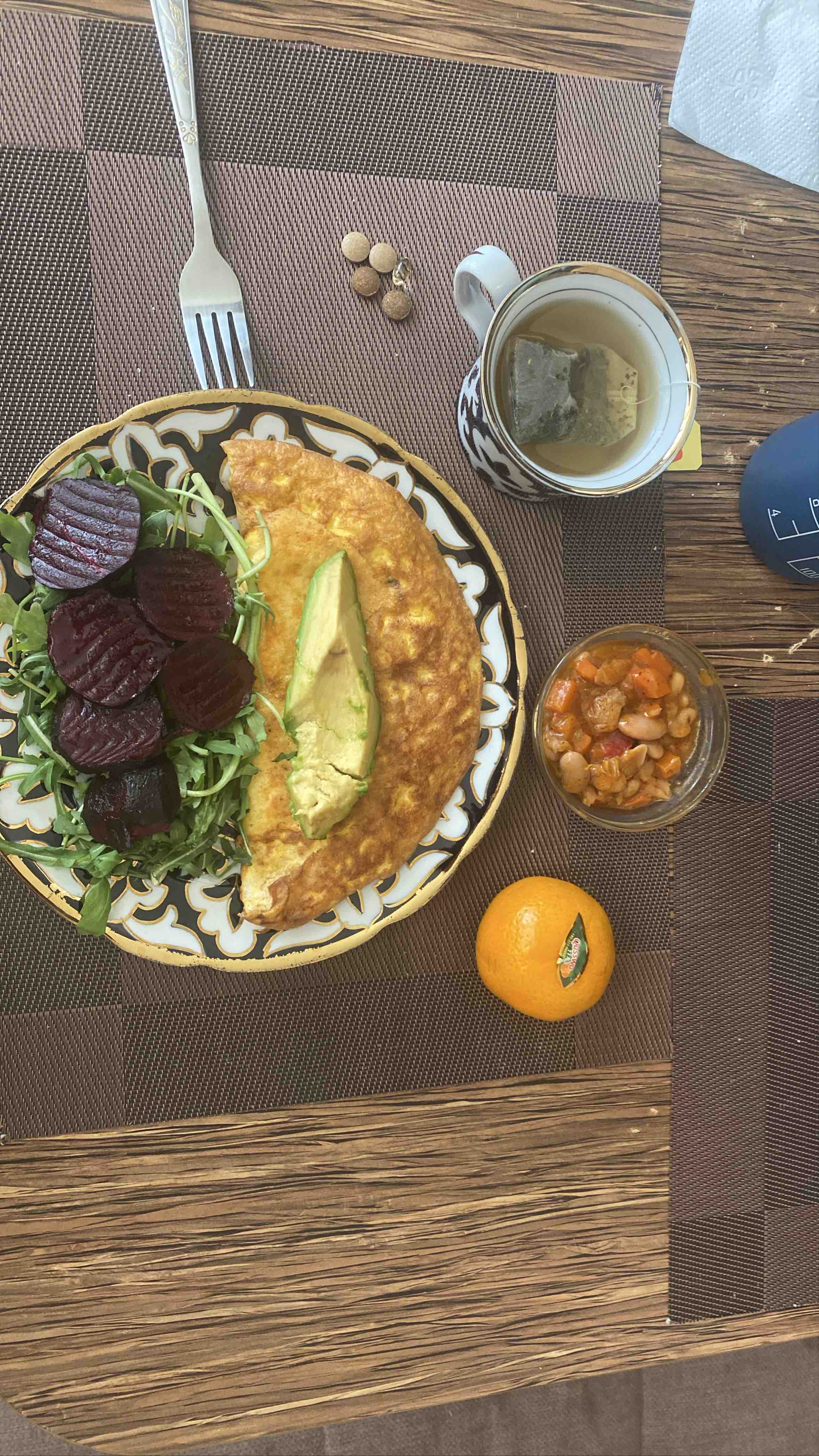 A balanced meal consisting of an omelette with low-fat cheese and turkey ham, avocado, beetroot, mixed greens, a side of bean stew, and a mandarin orange, accompanied by herbal tea.