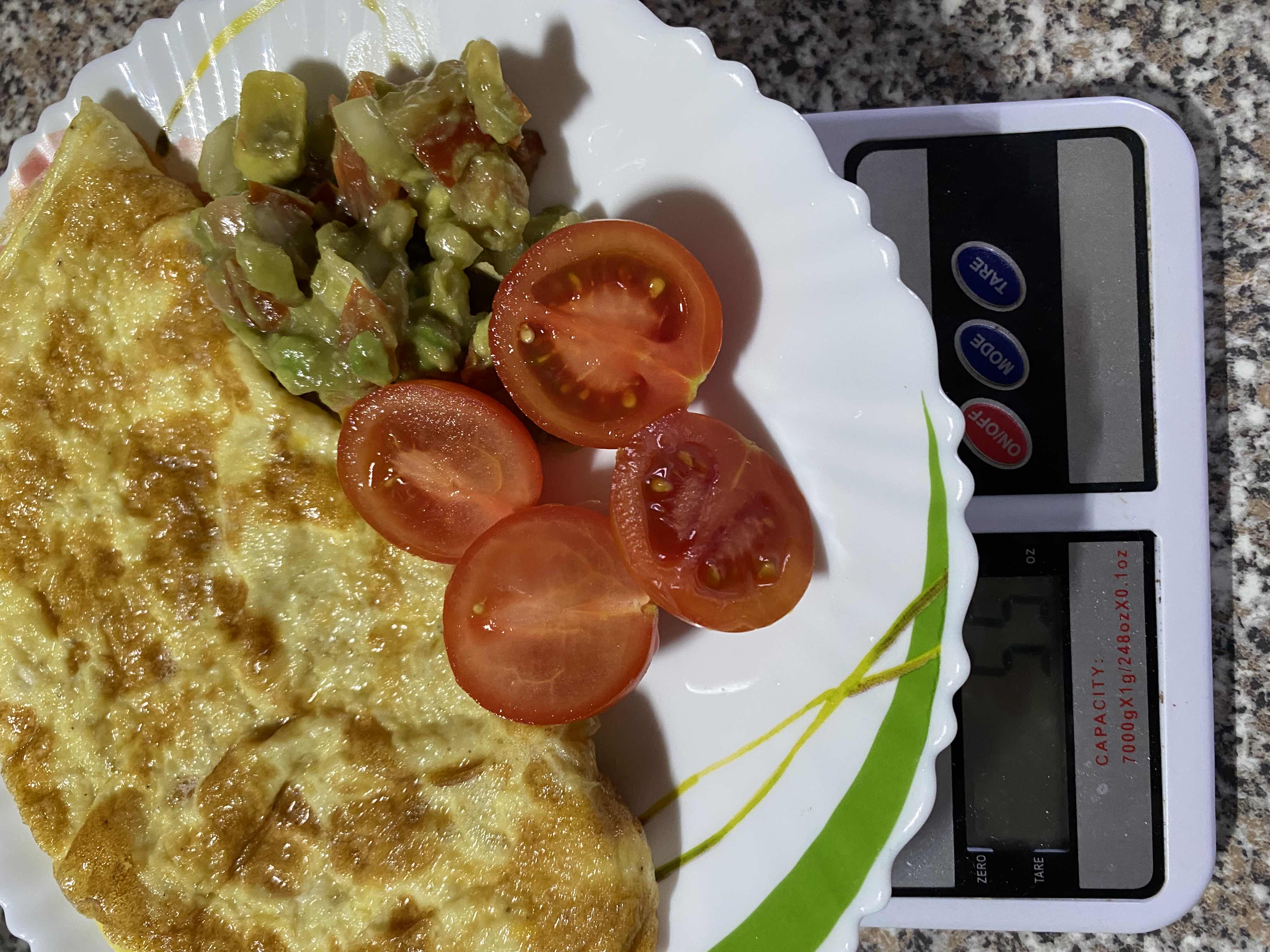 A plate containing an omelette, sliced tomatoes, and an avocado salad, placed on a digital scale showing a weight of 55 grams.