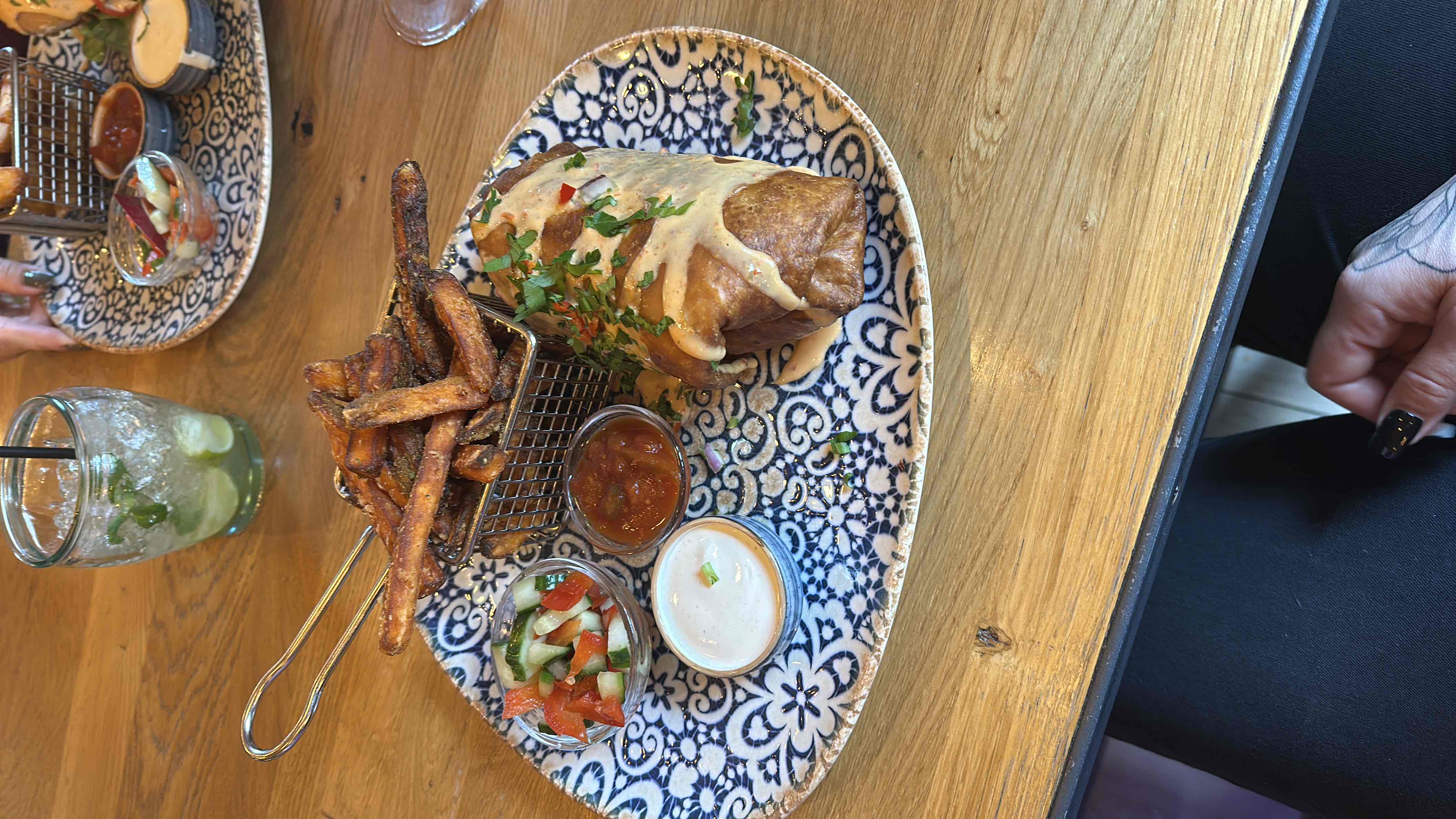 A plate featuring a burrito topped with sauce, sweet potato fries, and small servings of salsa and cream sauce.