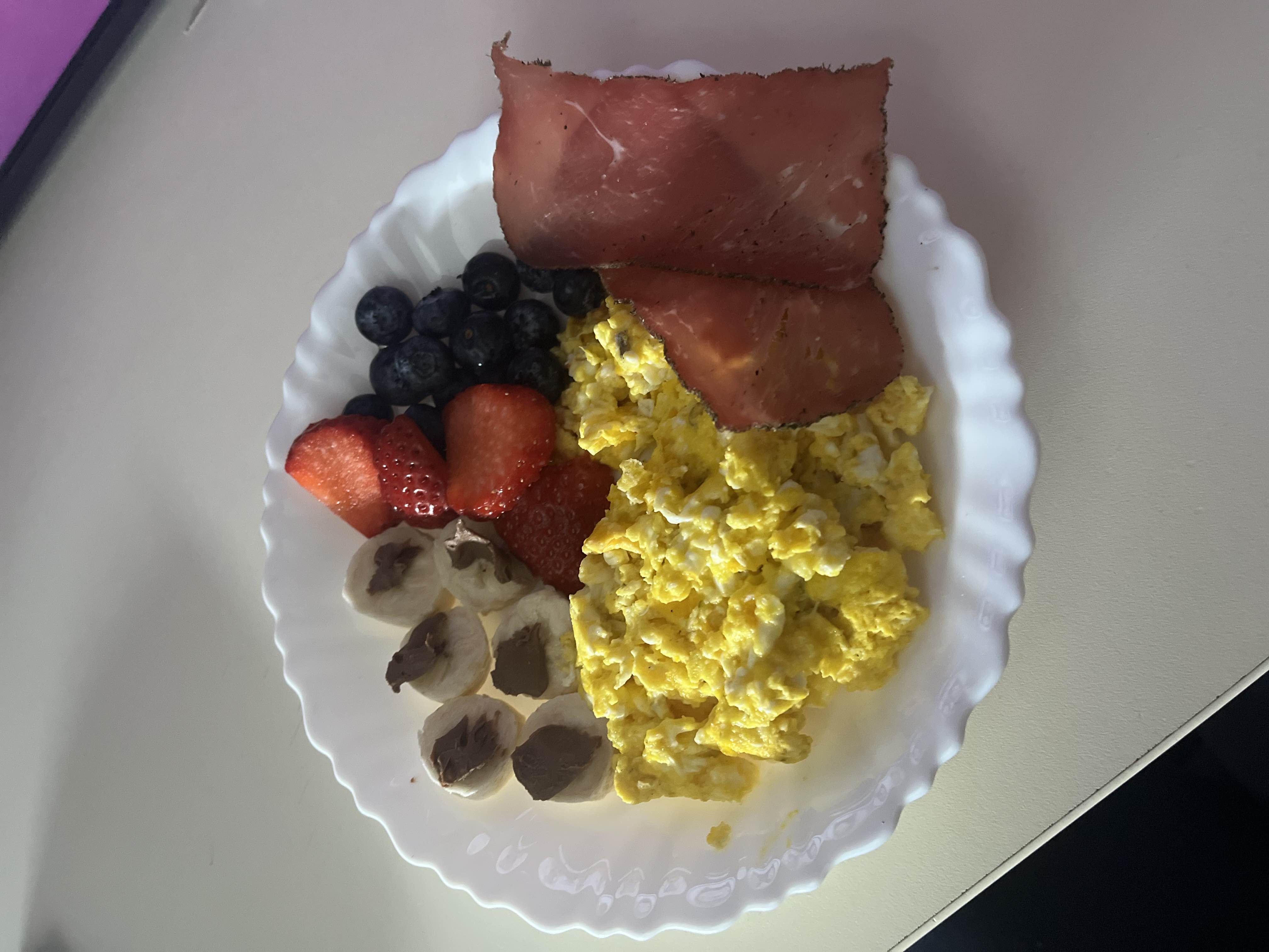 A balanced plate featuring scrambled eggs, fresh fruits (strawberries, blueberries, and banana slices with chocolate spread), and slices of cured meat.