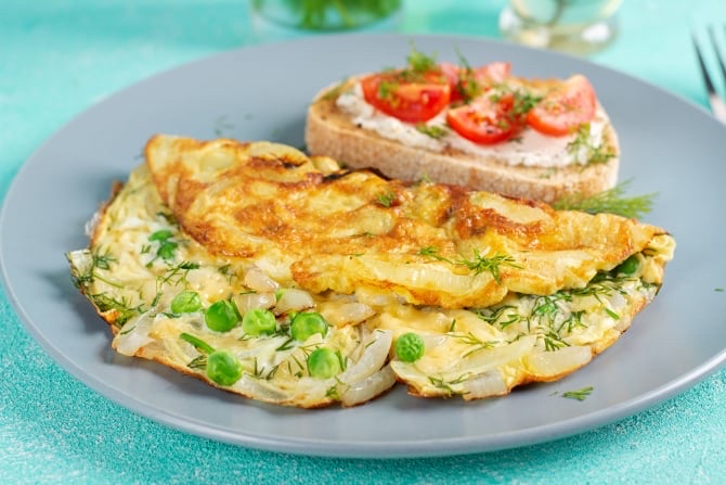 A nutritious meal featuring an omelette with peas and herbs, served alongside a slice of bread topped with cream cheese and fresh tomatoes.