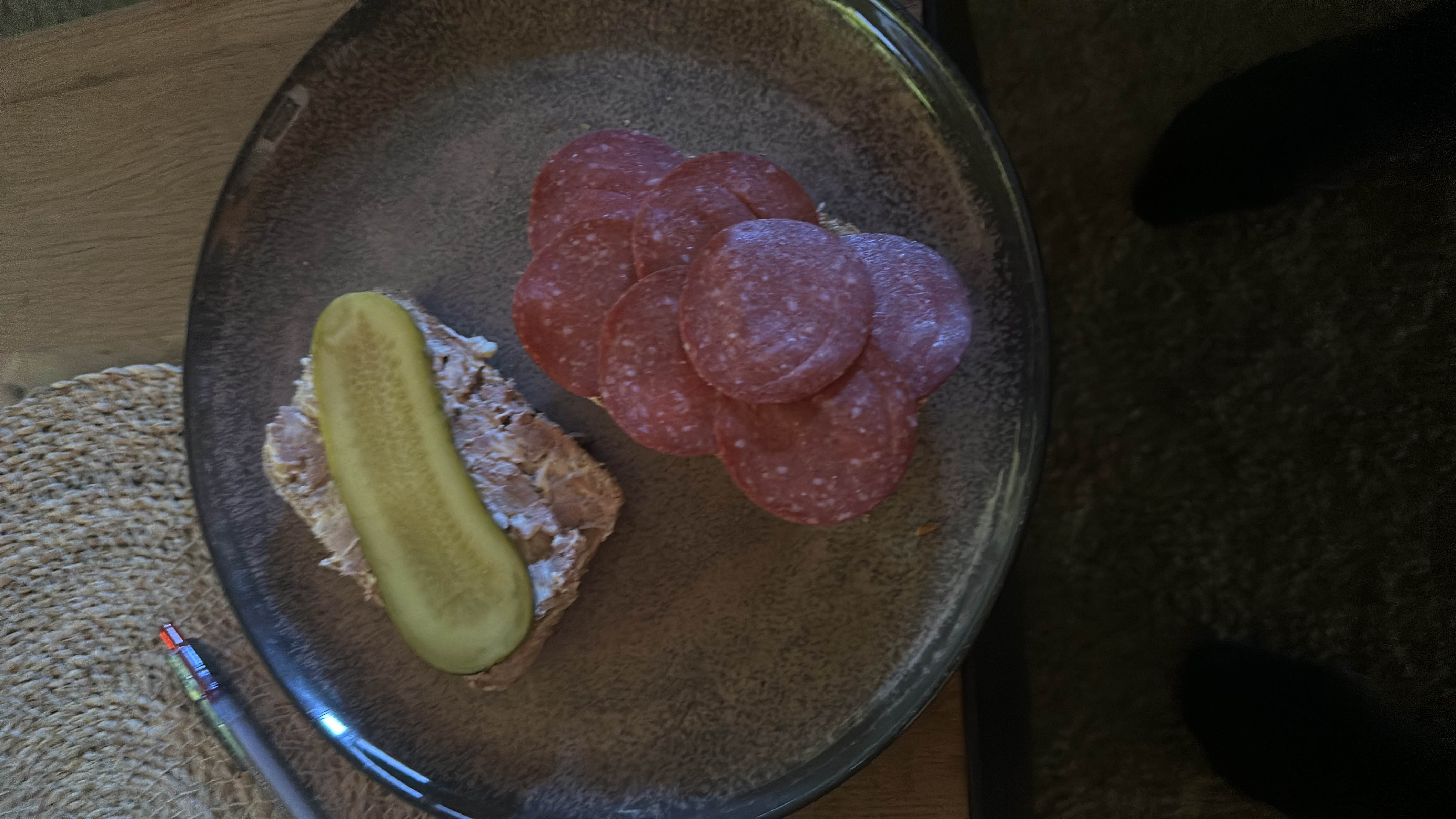 A plate containing salami slices, a piece of bread with spread, and a pickle slice, providing a balanced mix of protein, fats, and carbs.