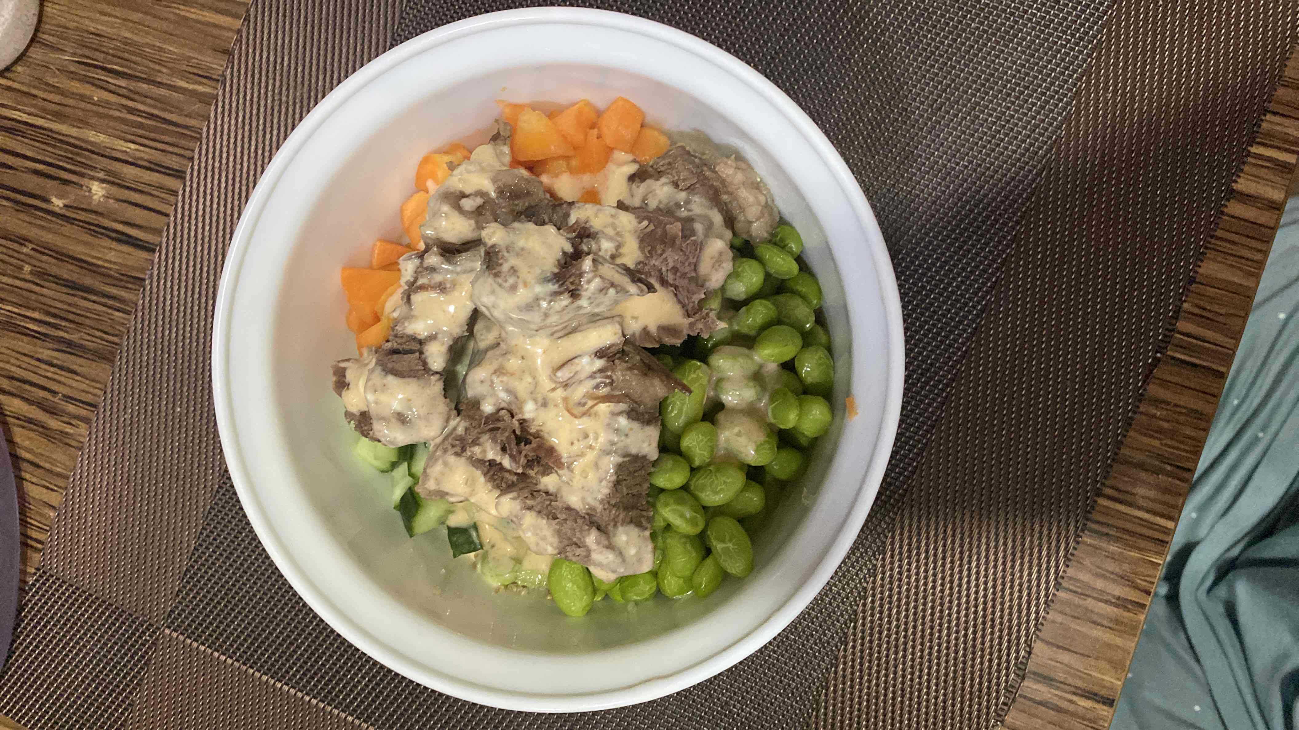 A balanced bowl containing beef, edamame, cucumber, carrot, creamy dressing, quinoa, and avocado.
