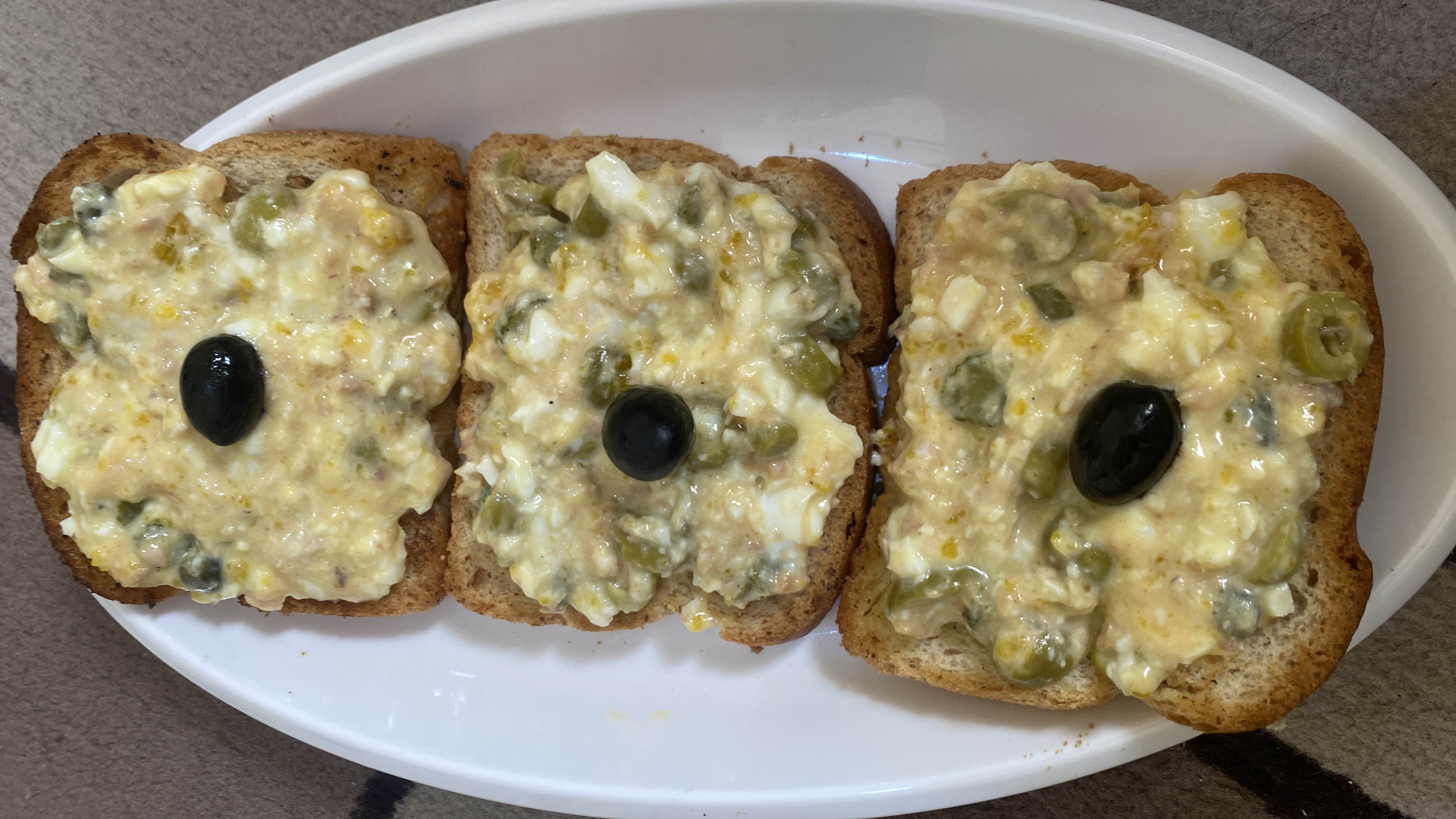 Three slices of bread topped with egg salad, green olives, and a single black olive on each slice.