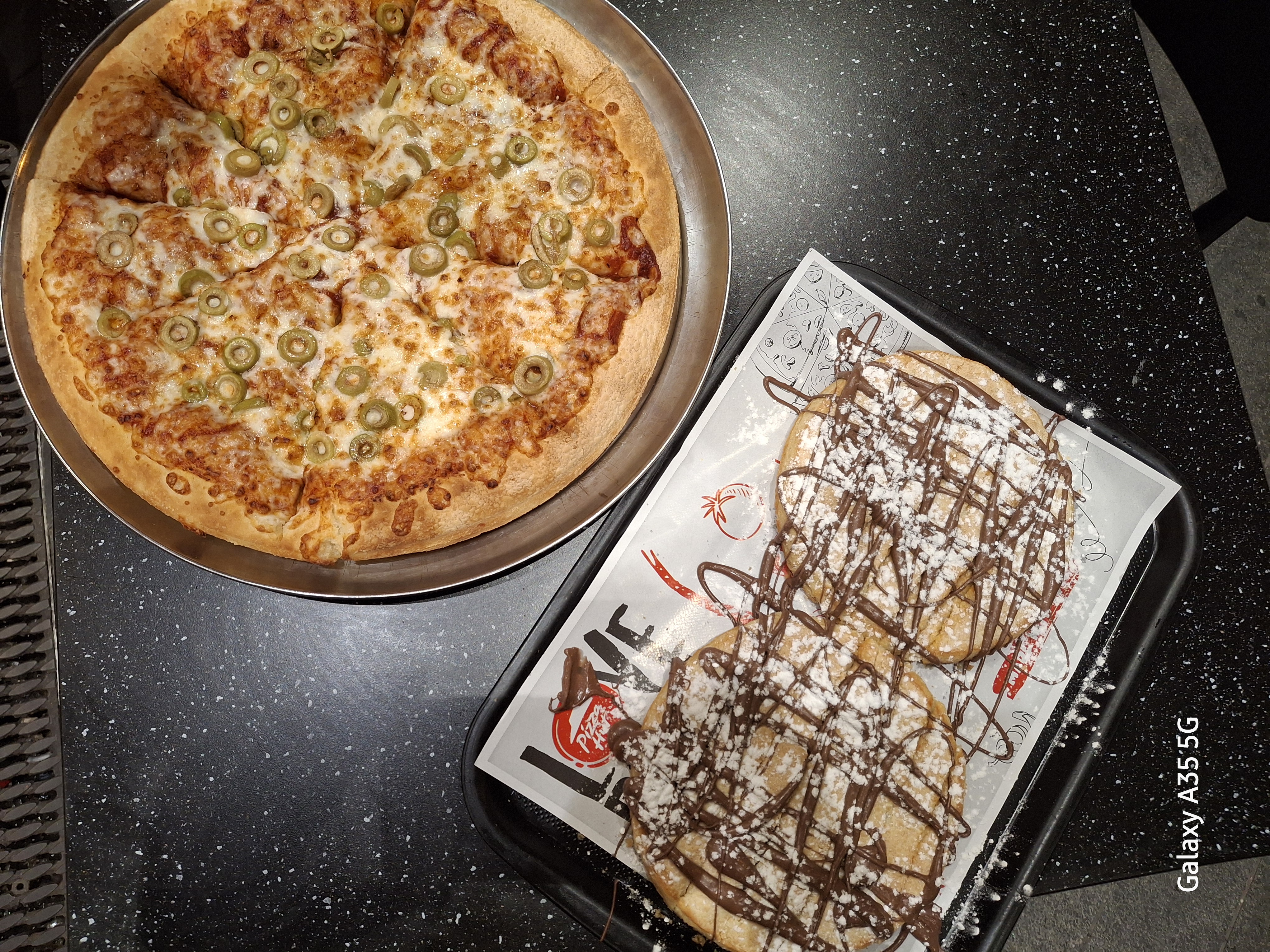 The image shows a pizza topped with olives and cheese alongside chocolate-drizzled pancakes, offering a mix of savory and sweet dishes.