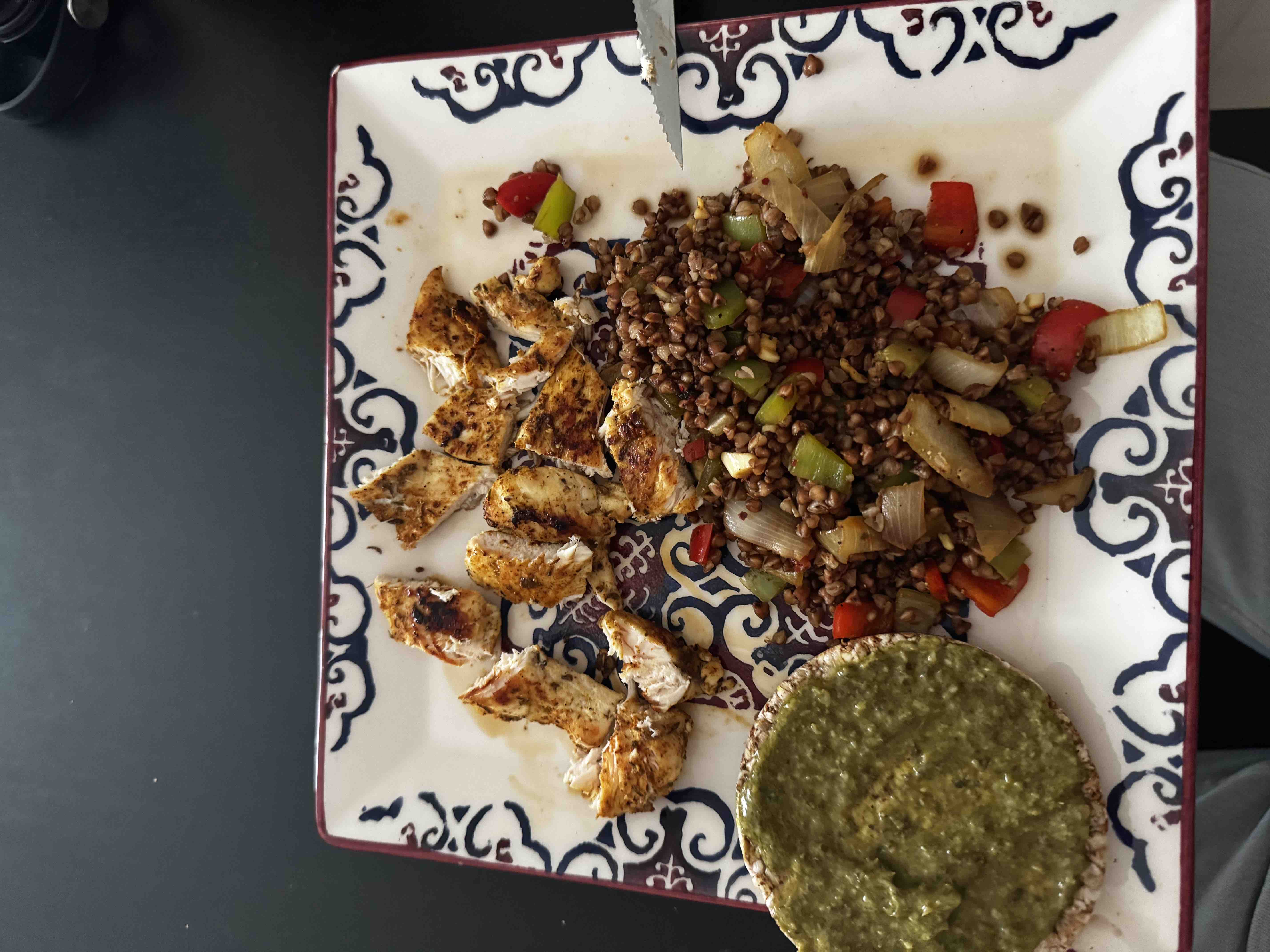 A balanced meal consisting of grilled chicken, buckwheat salad with vegetables, and a rice cake topped with pesto.