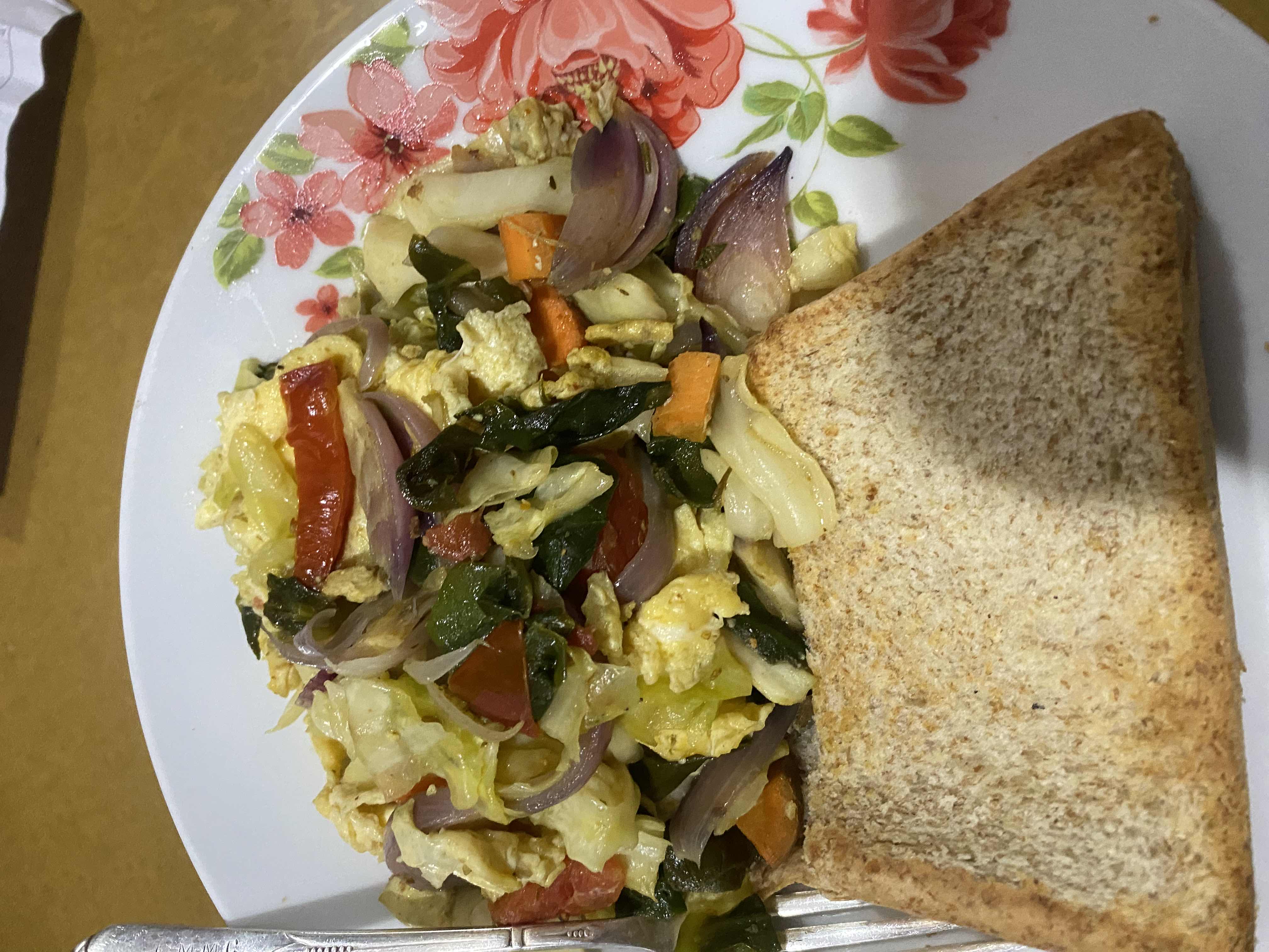 A healthy meal consisting of scrambled eggs mixed with sautéed vegetables such as onions, tomatoes, carrots, cabbage, and spinach, served with a slice of whole wheat bread.