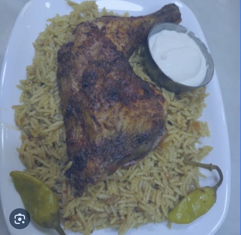 A plate of spiced rice served with a grilled chicken leg, yogurt sauce, and green chili peppers, providing a balanced mix of protein, carbs, and fats.