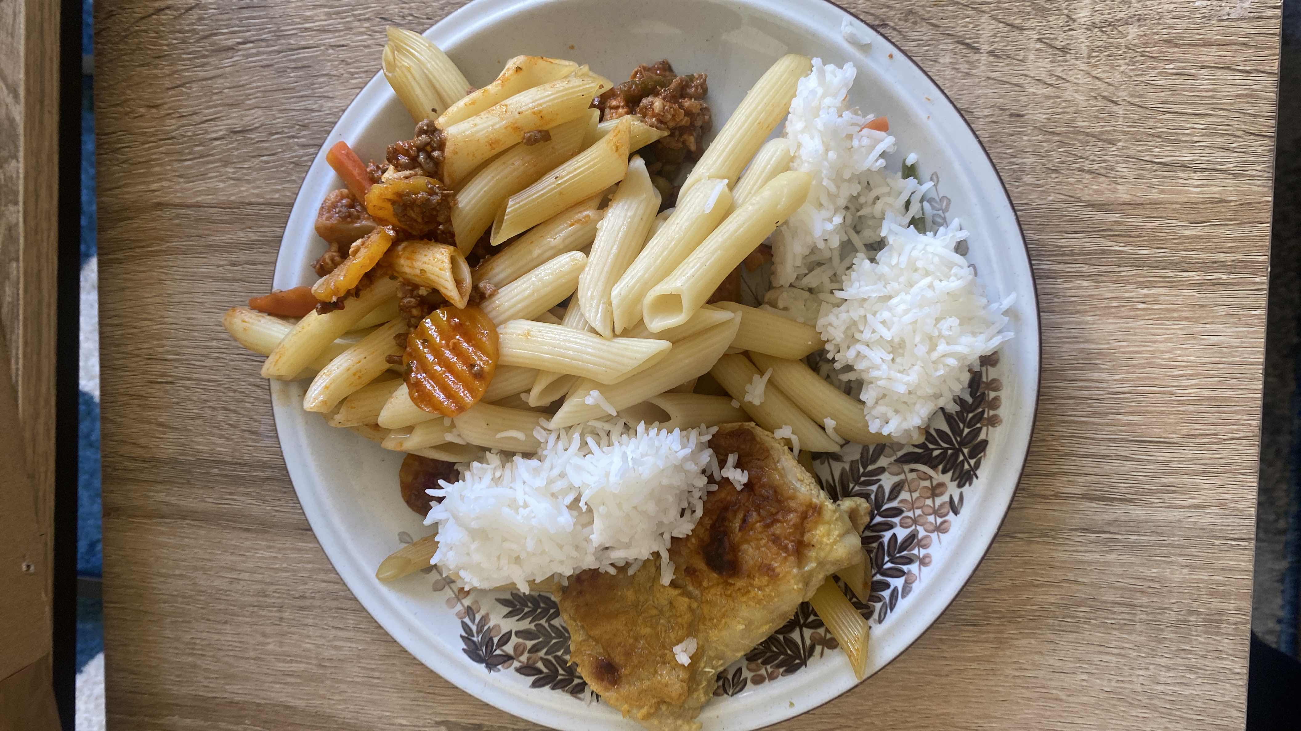 A plate containing pasta with meat sauce, white rice, and a piece of baked or fried chicken, accompanied by vegetables.