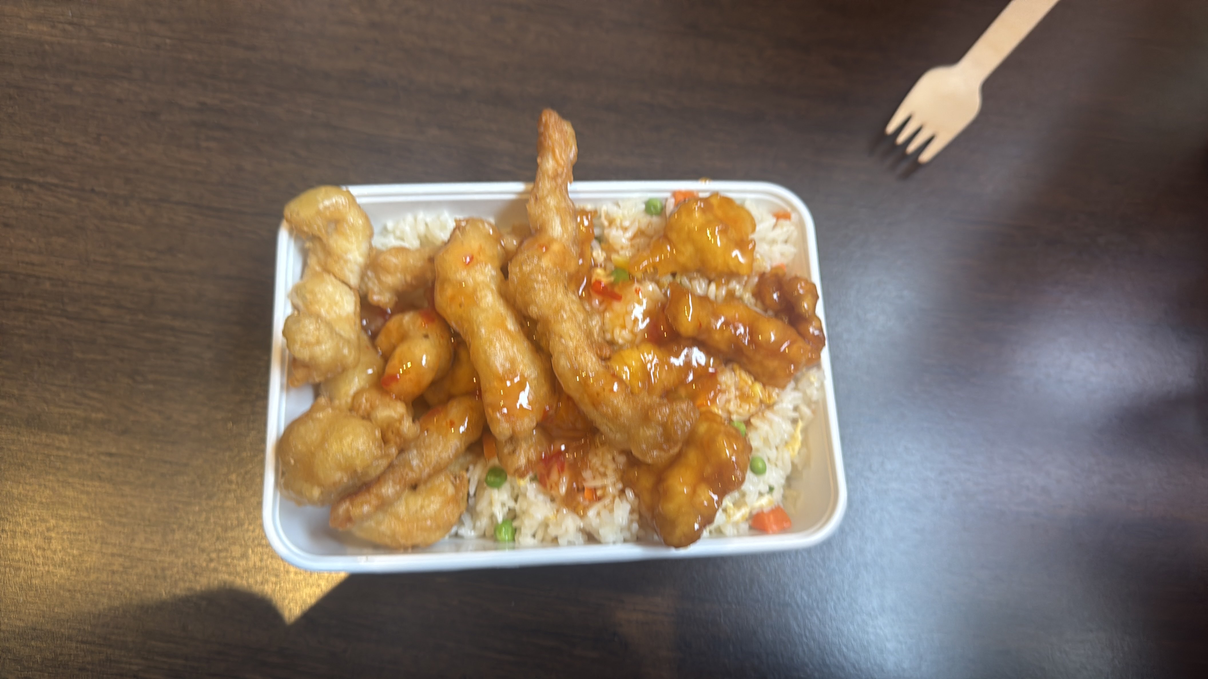 A takeout container filled with fried rice, topped with battered and fried chicken strips coated in a sweet chili sauce.