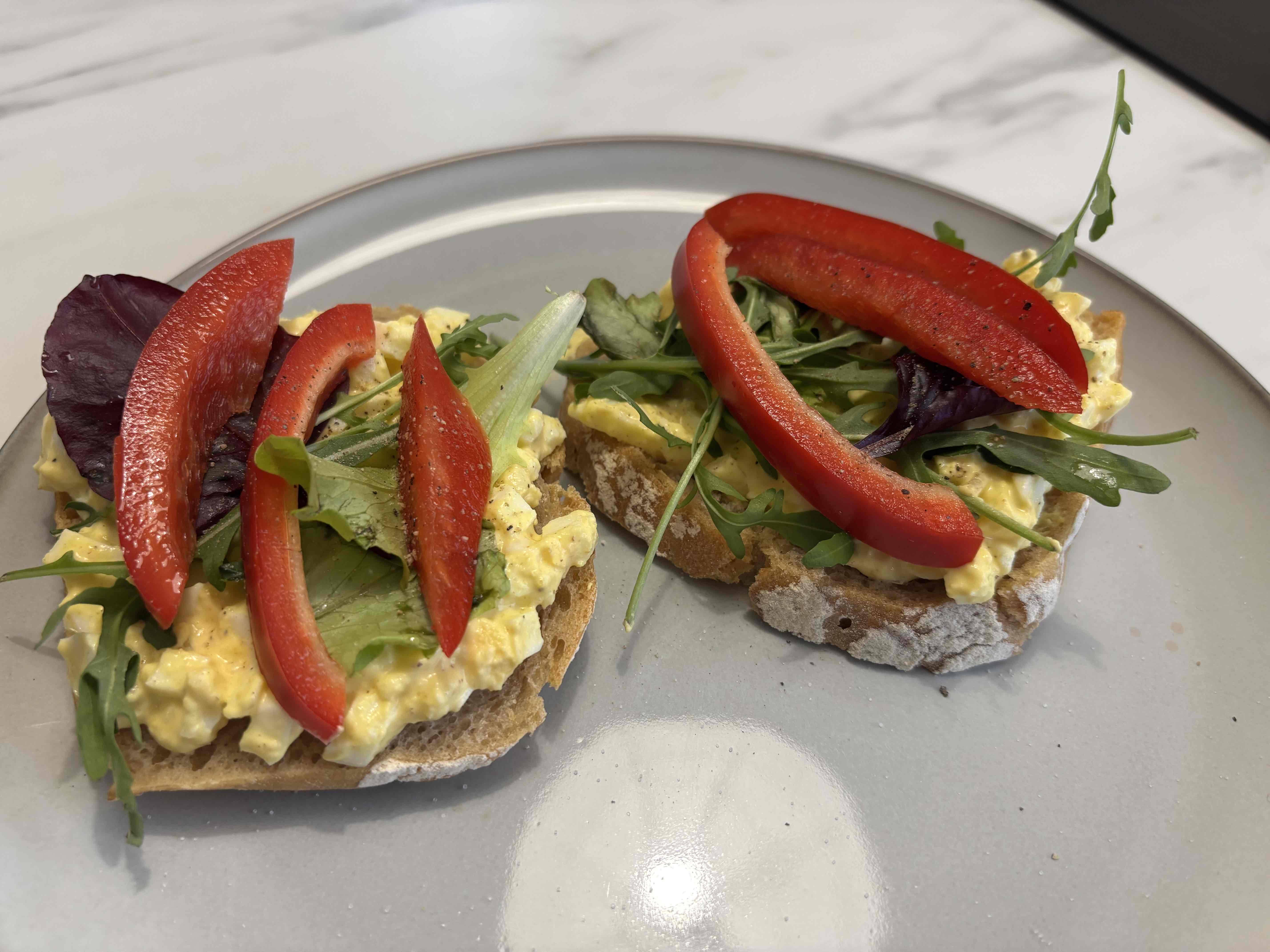 Open-faced sandwich with egg salad, red bell pepper slices, and mixed greens on bread.