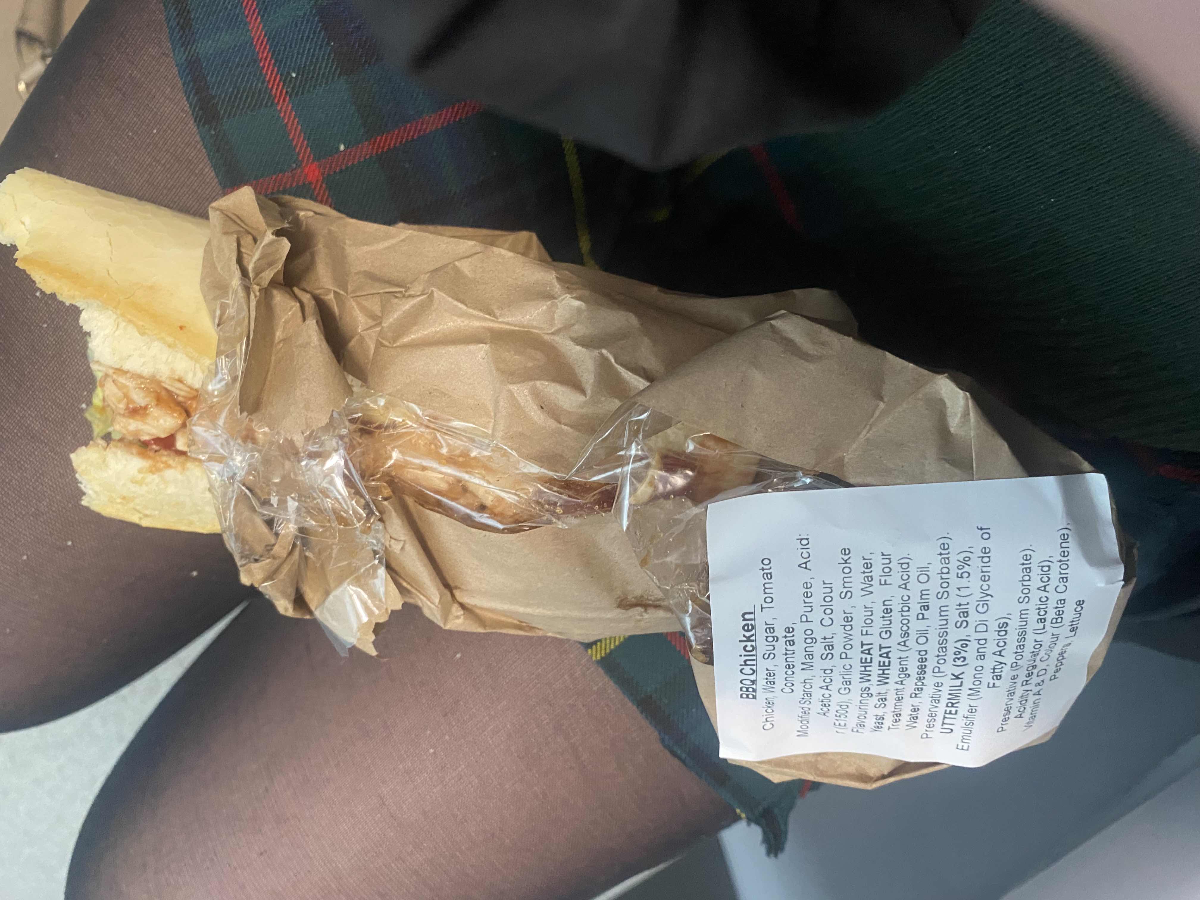 A BBQ chicken sandwich with bread, chicken, and BBQ sauce, wrapped in paper packaging.