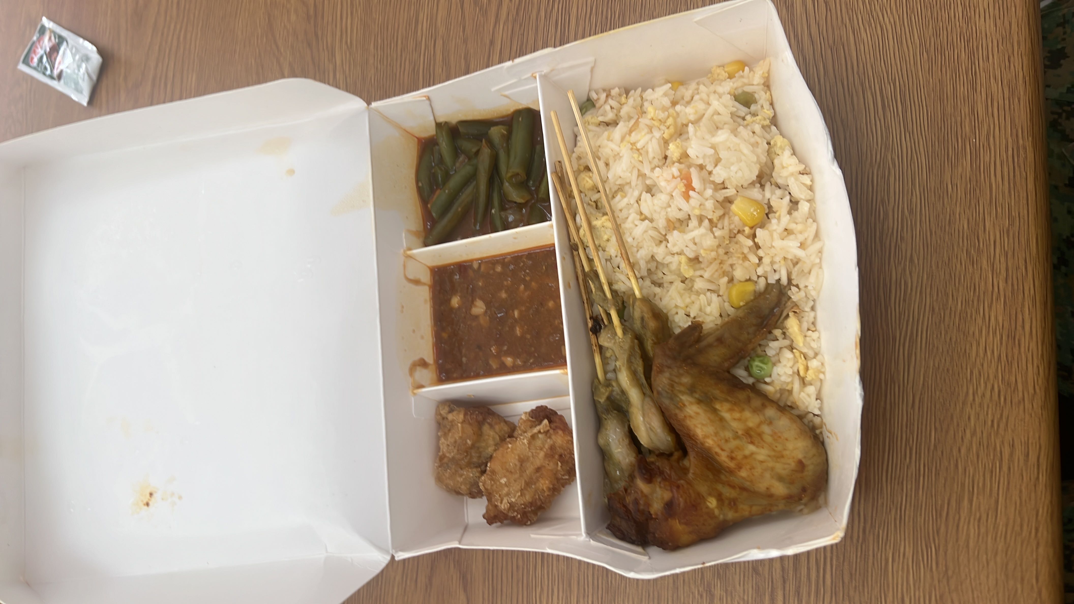 A meal box containing fried rice with mixed vegetables, grilled chicken wing, chicken satay skewers, fried chicken, green beans in sauce, and a side of peanut sauce.