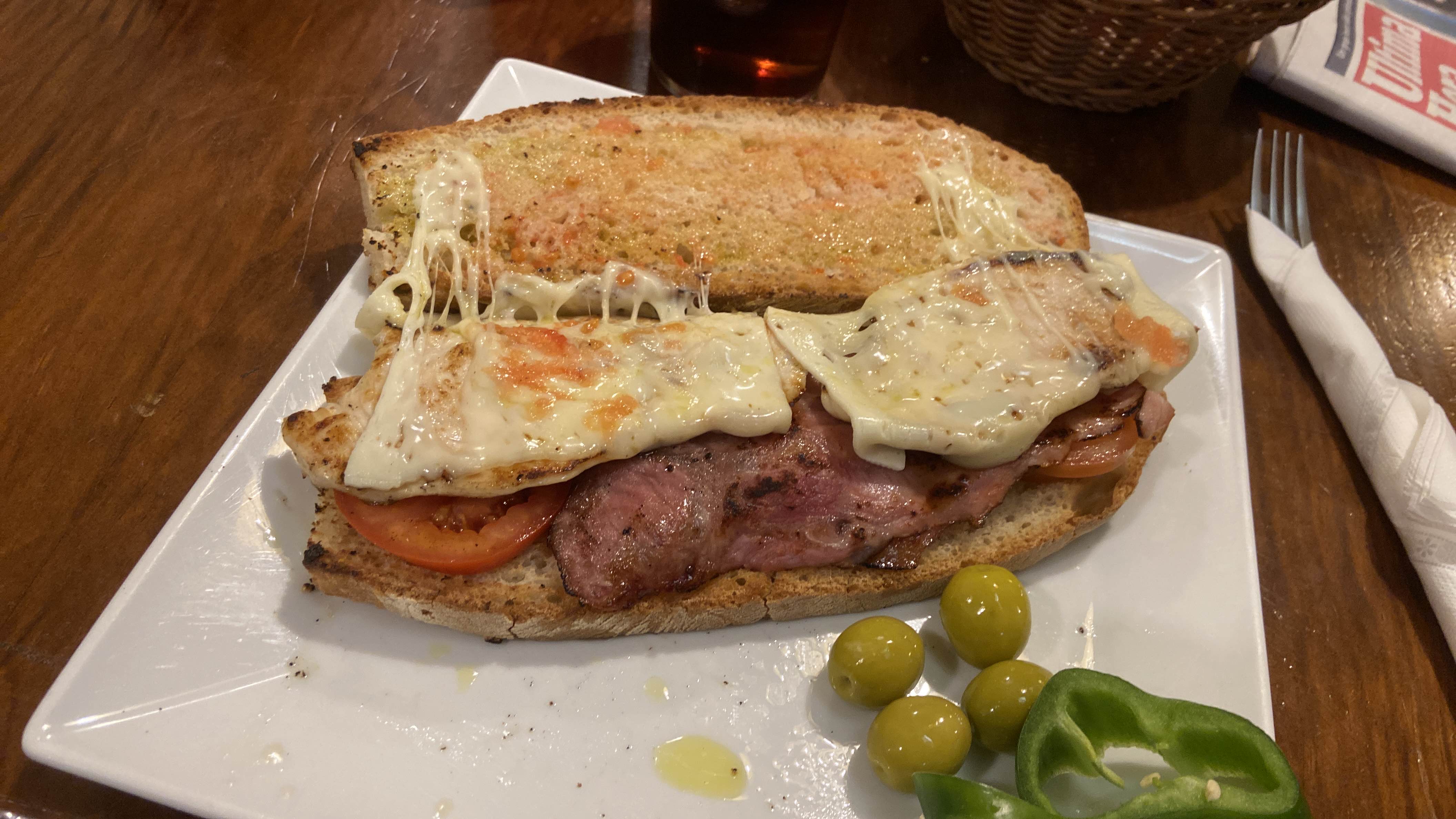 A toasted sandwich with grilled chicken, bacon, melted cheese, and tomato, served with green olives and bell pepper slices.