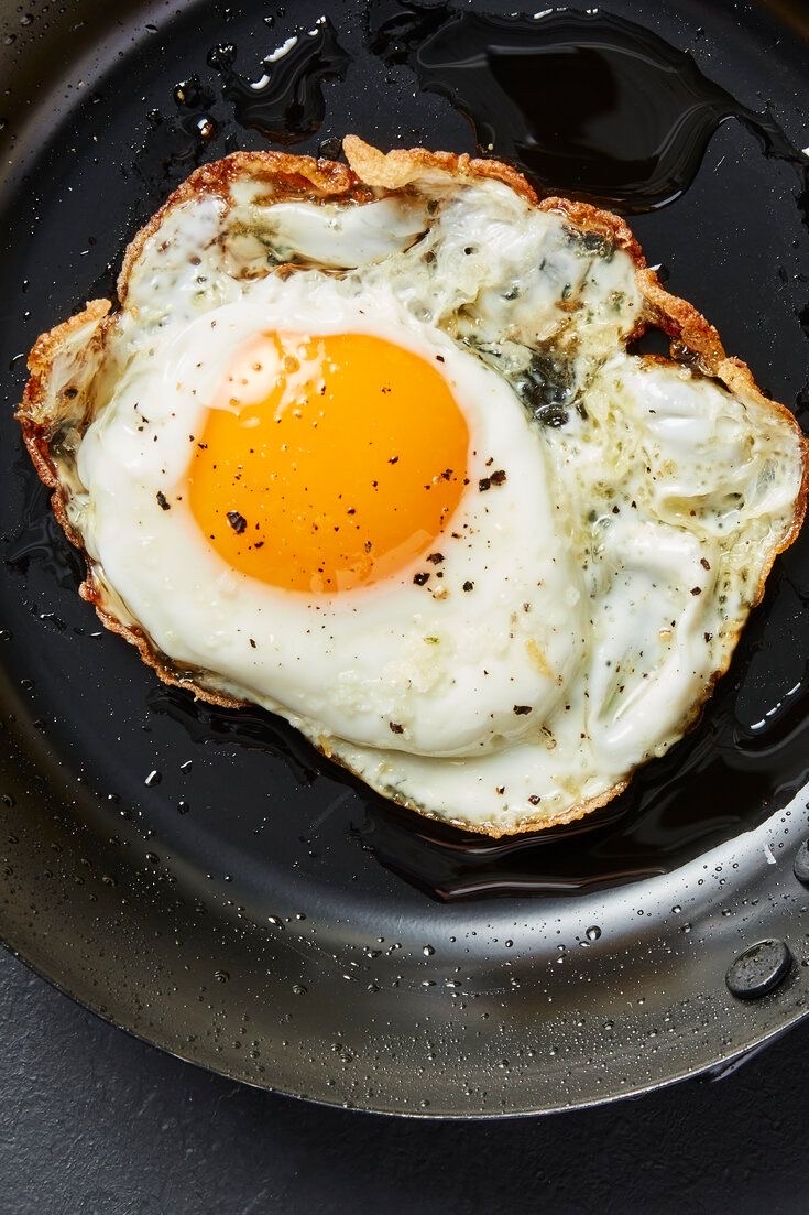 A fried egg with crispy edges, seasoned with black pepper, cooked in oil on a black pan.