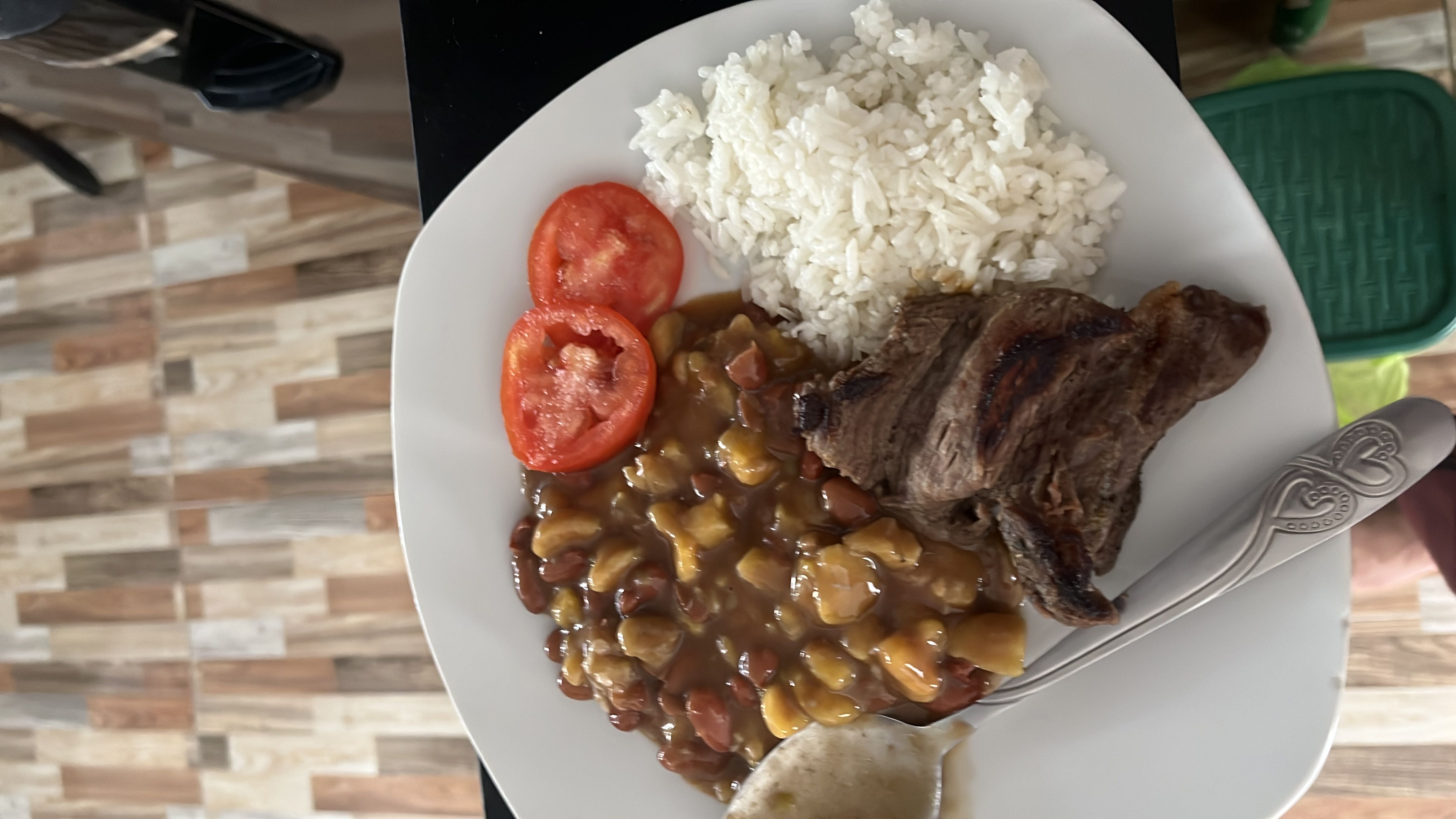 A balanced meal consisting of grilled beef, white rice, beans with plantains, and fresh tomato slices, providing a good mix of protein, carbohydrates, and fats.