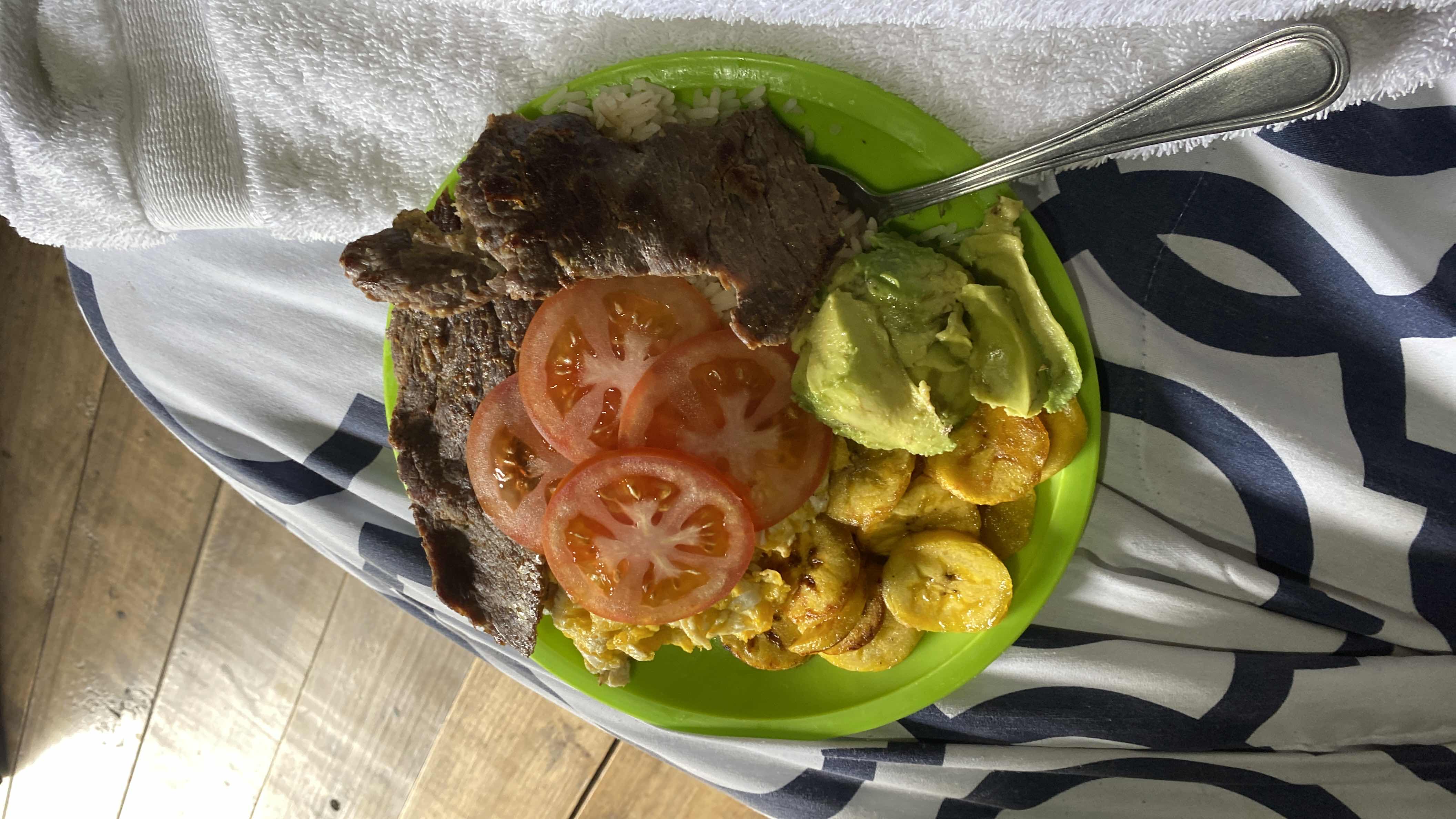 A balanced meal consisting of grilled beef, white rice, fried plantains, scrambled eggs, fresh tomato slices, and avocado, providing a mix of protein, carbohydrates, and healthy fats.