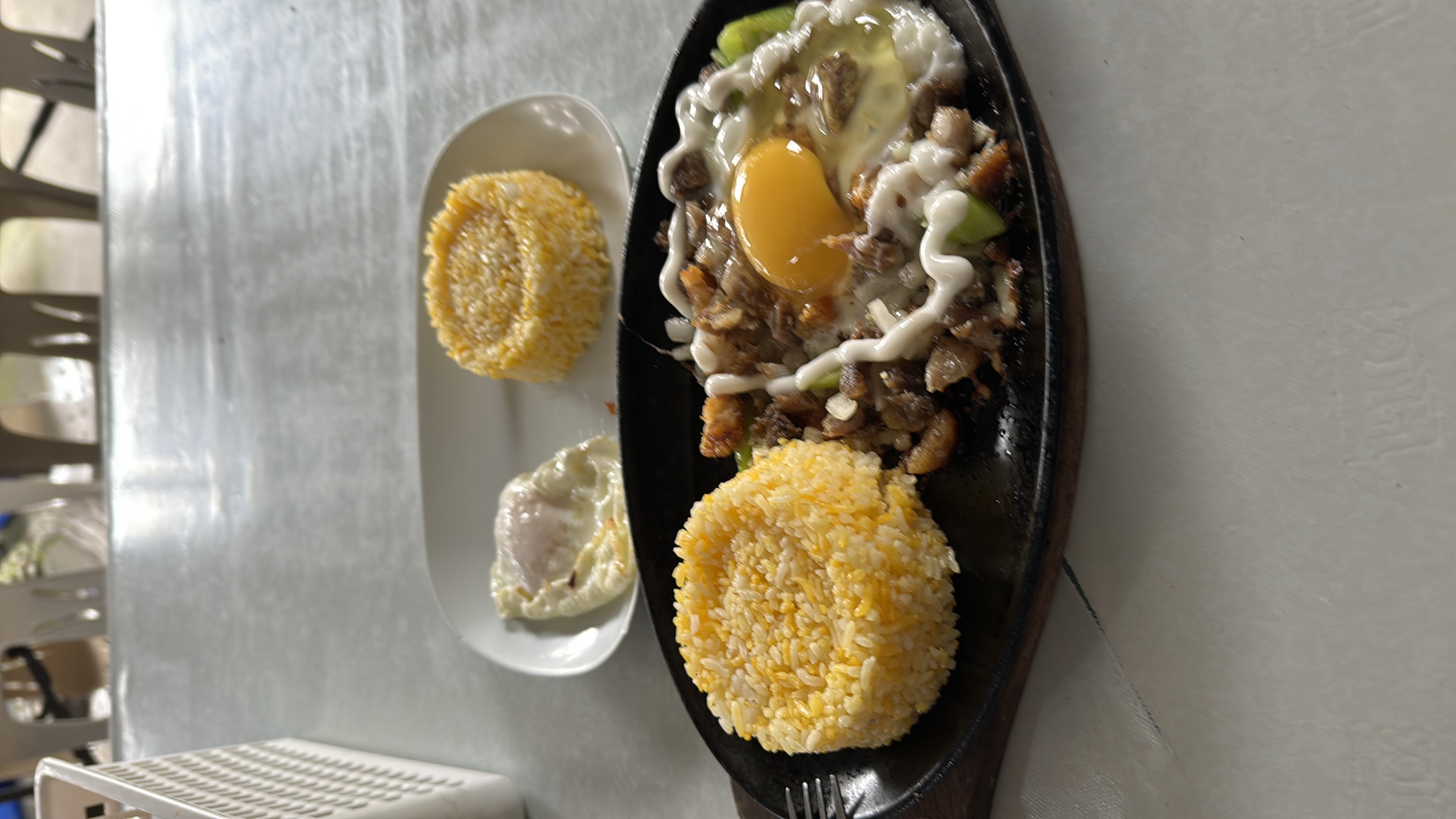 A Filipino meal consisting of garlic rice, pork sisig topped with a raw egg, and a side of fried egg, served on a sizzling plate.