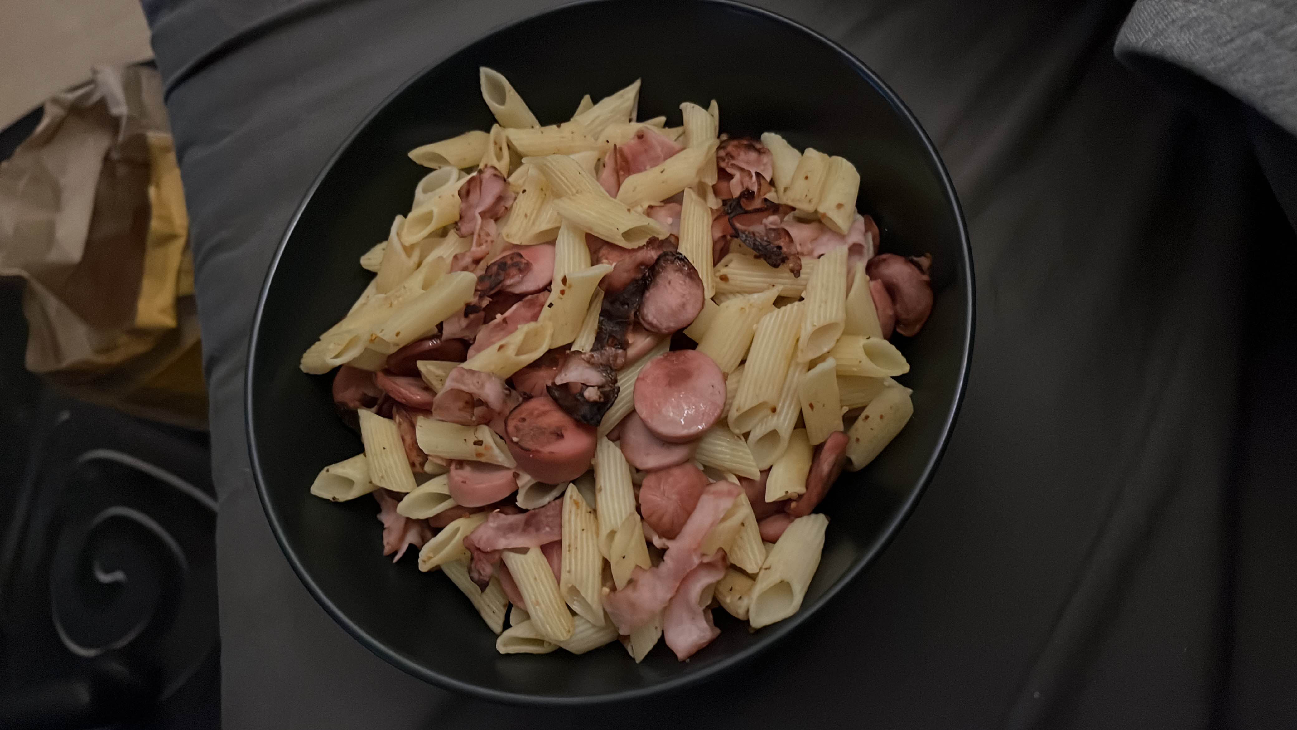 A bowl of penne pasta mixed with sliced sausages and ham, lightly seasoned with spices.